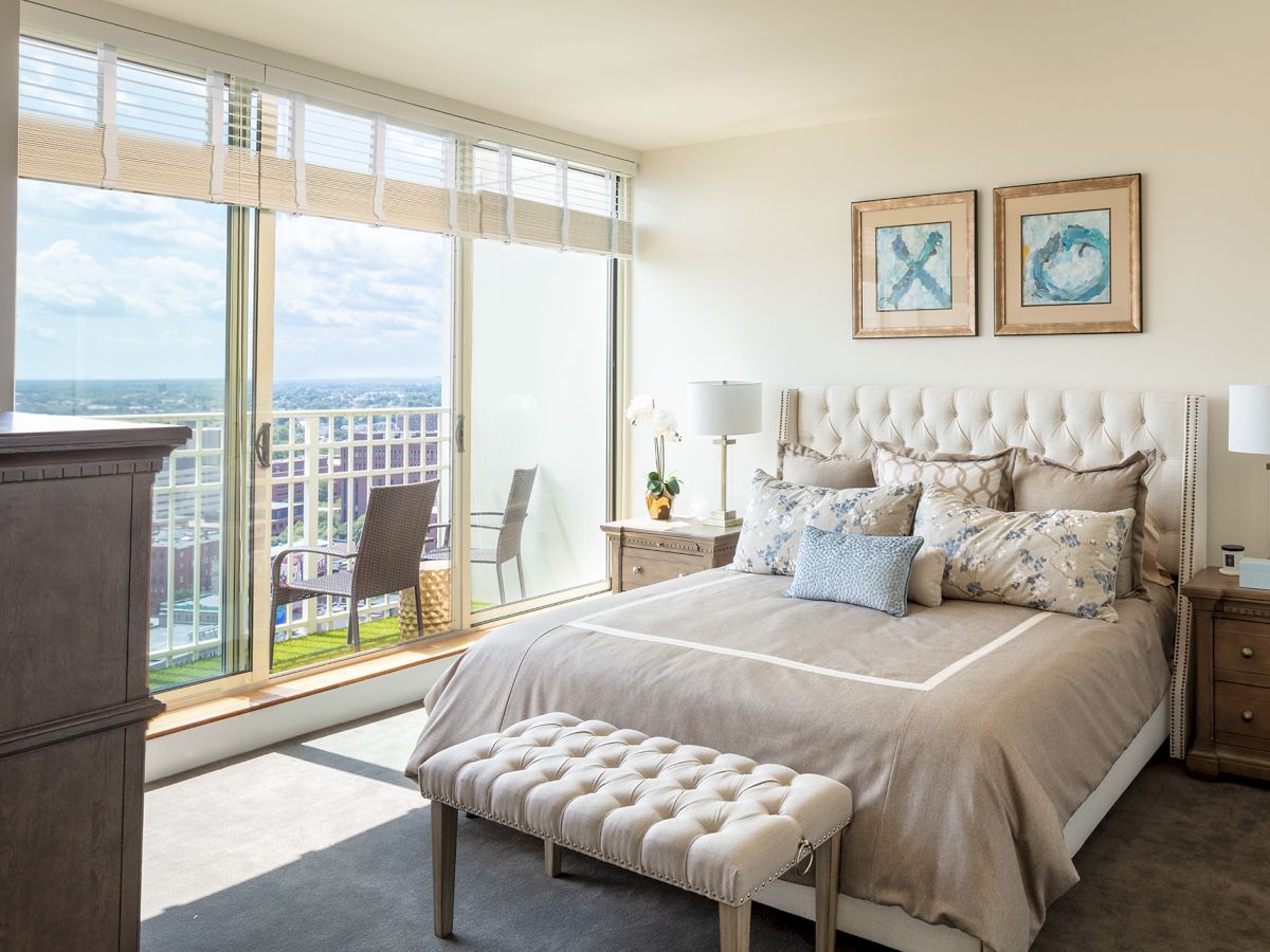 This image features a modern bedroom with a large window offering a city view, a cushioned bed, side tables with lamps, and framed wall art.