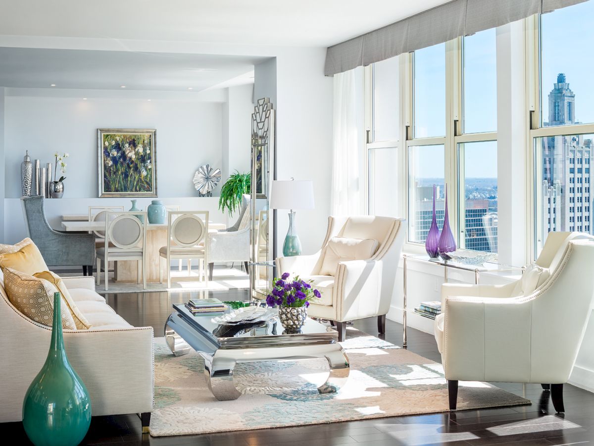 The image shows a modern, well-lit living and dining area with large windows, contemporary furniture, and city views, including a tall building outside.