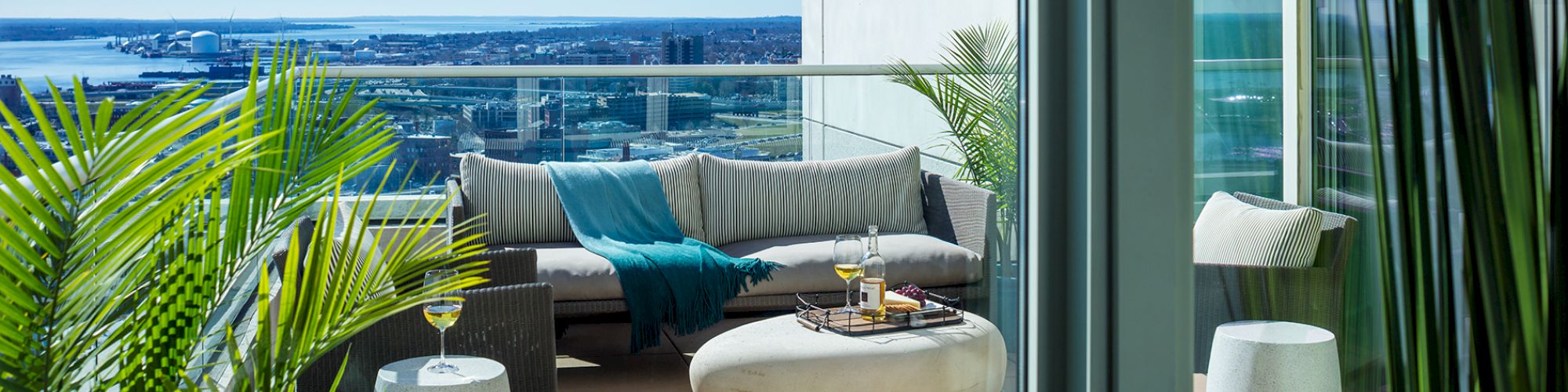 A balcony with modern furniture, including a sofa and round table, overlooking a distant body of water. Various potted plants are present ending the sentence.