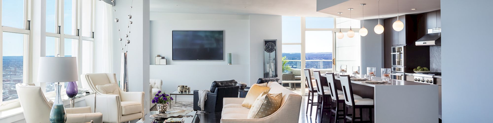 A modern open-concept living room and kitchen with large windows, a TV, white sofas, a coffee table, and a dining area with pendant lights.