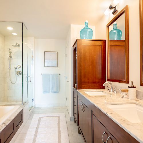 This image shows a modern bathroom with a shower, bathtub, double sink vanity with mirrors, wooden cabinets, and blue decorative vases ending the sentence.