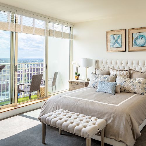 A modern bedroom features a plush bed with multiple pillows, a bench at its foot, large windows with a view, and two pieces of wall art.