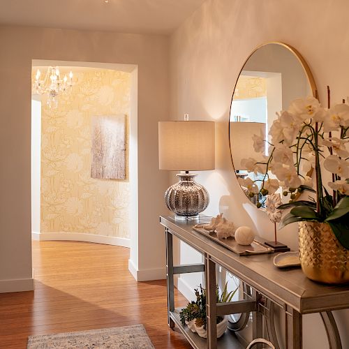 A stylish hallway features a console table with a lamp, mirror, and orchid, leading to another room with hardwood flooring and a chandelier.