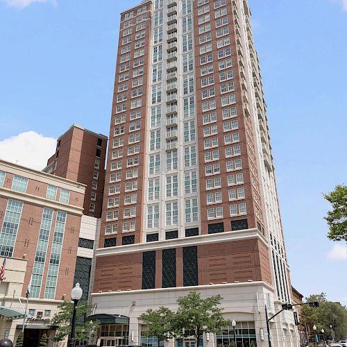 A tall, multi-story building with a mix of red brick and white facade stands against a partly cloudy sky, next to a streetlamp and trees.