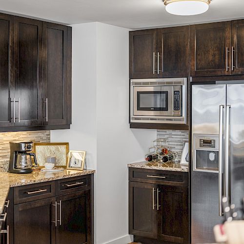 A modern kitchen with dark cabinets, a stainless steel fridge, a microwave, a coffeemaker, a backsplash, and granite countertops, with a few decor items.