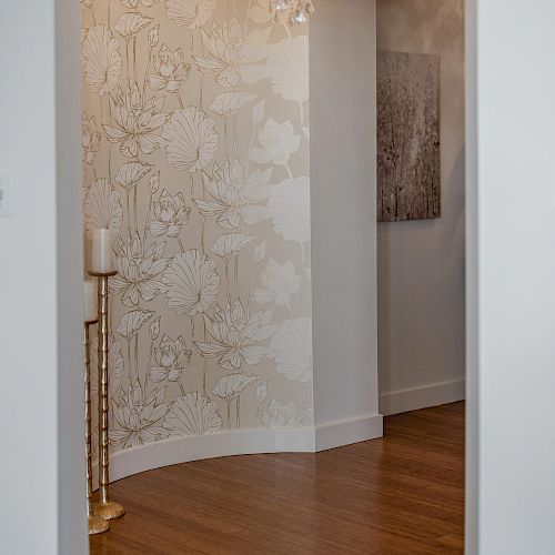 A hallway with wood flooring, floral wallpaper, a chandelier, a tall gold candle holder, and a piece of artwork on the wall.