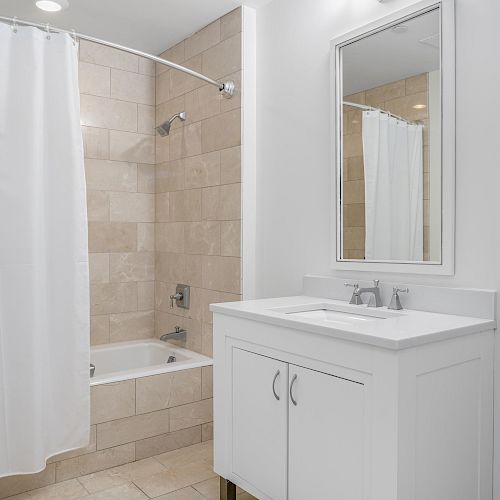 This image shows a clean, modern bathroom with a white vanity and mirror, a bathtub with a shower curtain, and beige tile walls and floors.