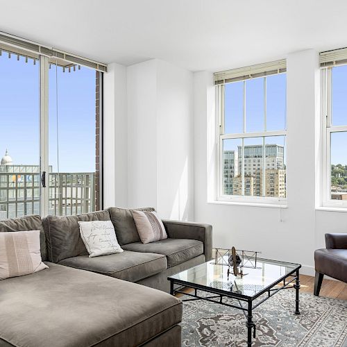 A modern living room with a sectional sofa, a brown armchair, a glass coffee table, and large windows with city views, ending the sentence.