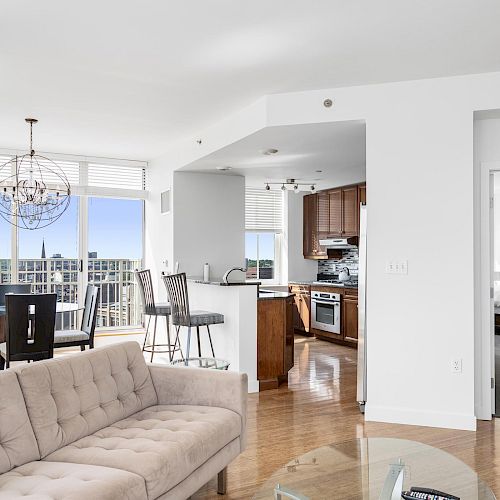 The image shows a modern apartment interior featuring a living room, dining area, kitchen, and a glimpse into a bedroom, decorated in neutral tones.