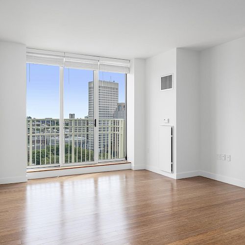 A bright, empty room with wooden floors, large windows, and a view of buildings outside. White walls and ceiling create a clean, modern look.