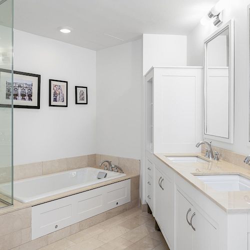 This image shows a modern bathroom with a bathtub, double sinks, mirrors, framed pictures on the wall, white cabinetry, and beige tile flooring.