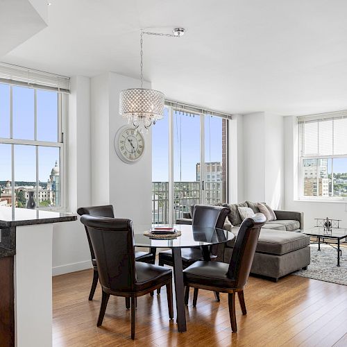 This image shows a modern living room with large windows, a dining table, chairs, sofa, and kitchen area in the background, all with bright natural light.