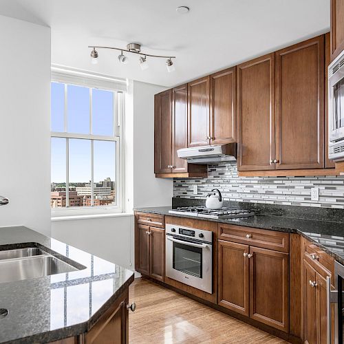 A modern kitchen with wooden cabinets, granite countertops, stainless steel appliances, a window, and a backsplash with a neutral pattern.