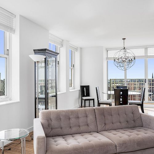 A modern living room with a plush sofa, glass tables, and a round dining table by the window. Large windows showcase a cityscape view.