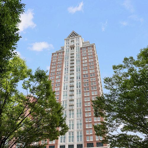 A tall building with a distinctive triangular top is framed by green trees, viewed from below on a clear day with a blue sky and some clouds.