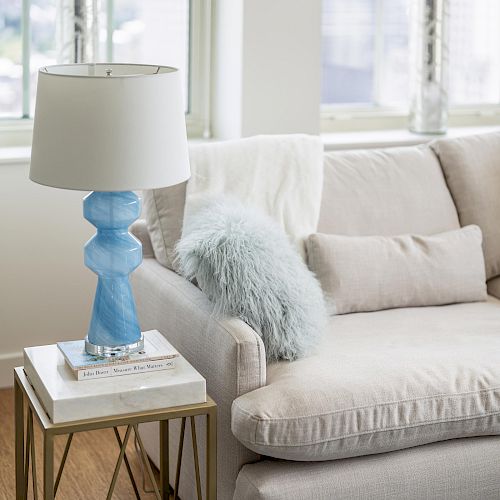 A cozy living room features a beige couch, a side table with a blue lamp and books, and a fluffy blue pillow, all bathed in natural light.