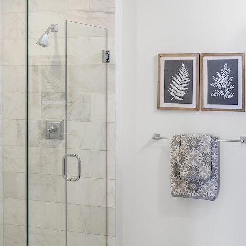 The image shows a bathroom with a glass-enclosed shower, two framed plant prints on the wall, and a towel hanging on a rail.