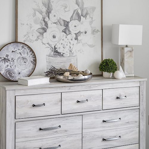 A light-colored dresser with multiple drawers is decorated with a floral painting, a table lamp, a tray, a plant, and a decorative plate.