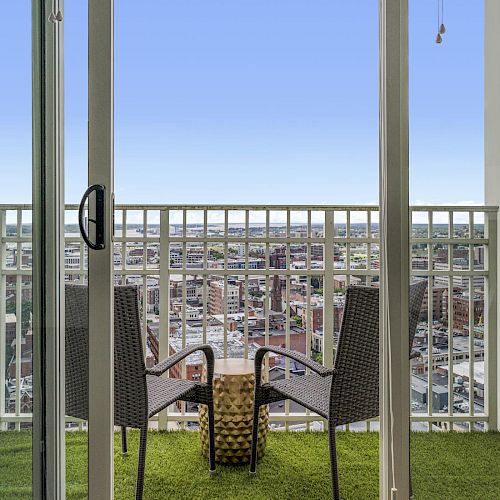 A balcony with artificial grass flooring, two wicker chairs, a small table, and a cityscape view through glass doors is depicted in the image.