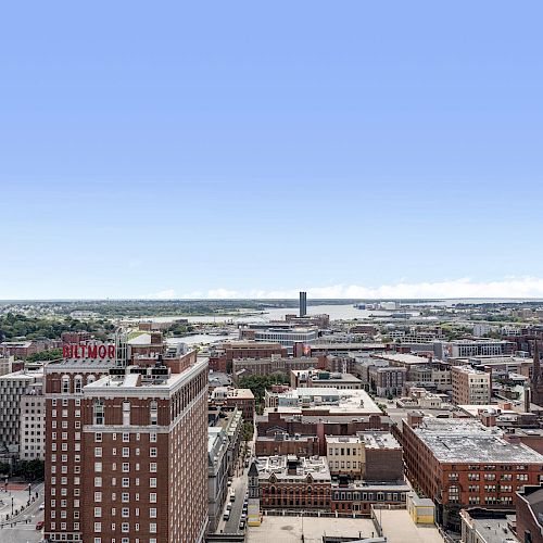 The image shows a city skyline with mid-rise buildings under a clear blue sky, with a prominent tall building on the left and a waterfront in the distance.