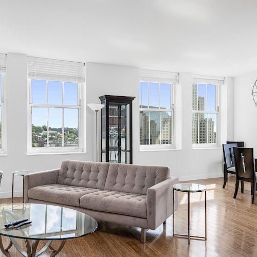 A modern living and dining area with large windows, a grey sofa, round glass coffee table, patterned armchair, dining table with chairs, and hardwood floors.