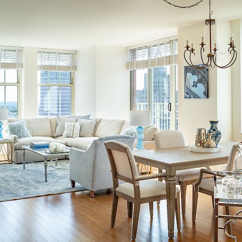 A bright, modern living and dining area with large windows, light furniture, blue accents, wooden floors, a chandelier, and city views is shown.