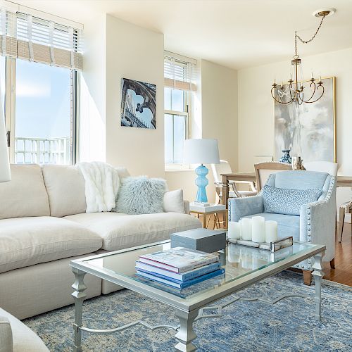 A modern living room features a white sofa, glass coffee table, blue rug, lamps, candles, and books, with a dining area in the background ending the sentence.