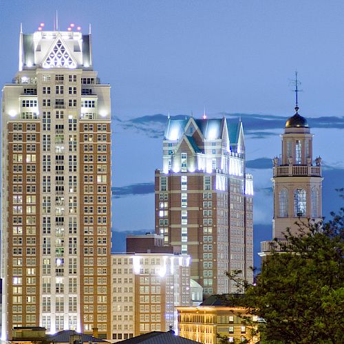 A cityscape at dusk featuring illuminated tall buildings with distinct architectural designs and a clear evening sky in the background.