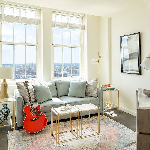 A bright living room with a large window, a blue sofa, a red guitar, decorative tables, lamps, wall art, and a rug on the floor, ending the sentence.