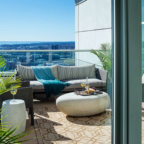 A cozy balcony with modern furniture, lush plants, and a stunning cityscape and water view in the background, featuring drinks on a table.