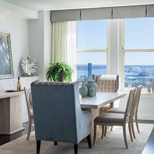 A modern dining room with a table, chairs, decorative vases, a large window, and a plant on the table, overlooking a picturesque outdoor view.