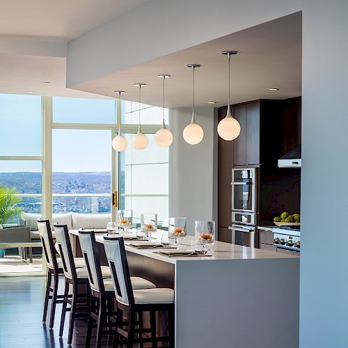 A modern kitchen with a dining area, featuring a long island with bar stools, overhead globe lights, and large windows with a view outside.