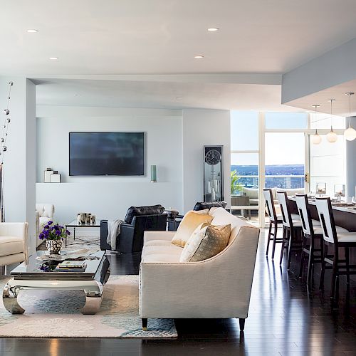 A modern open-plan living room with a kitchen/dining area. Features light furniture, large windows, and a wall-mounted TV.