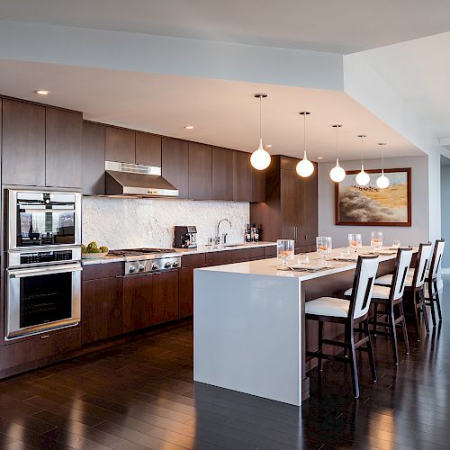 A modern kitchen with wooden cabinets, a large island, bar stools, pendant lights, and stainless steel appliances. Dining and living areas are visible.