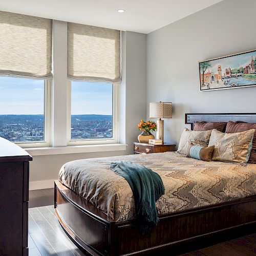 A modern bedroom with a large bed, two windows with beige shades, and a scenic view; a painting, lamps, and plants add to the decor.