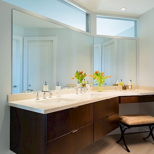 The image shows a modern bathroom with a large mirror, a floating vanity, sink, flowers, and a cushioned stool beneath the counter.