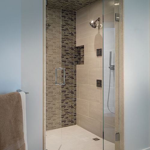 The image shows a modern glass-enclosed shower with tiled walls and a towel hanging outside on the left.