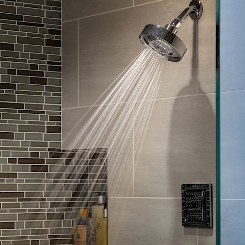 A modern shower with water spraying from the showerhead. The wall has a mix of mosaic and large tiles. There are toiletries on a shelf below.