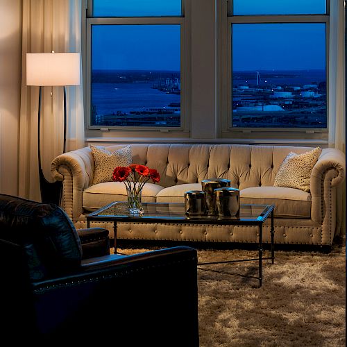 A cozy living room with a beige sofa, glass coffee table, and black armchair. There's a lamp, rug, and a window view of the city at dusk.