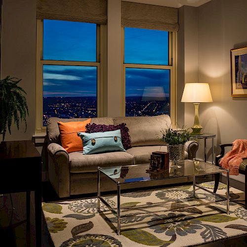 A cozy living room with a sofa, colorful pillows, a glass coffee table, and a floor lamp by the window, offering a stunning nighttime city view.