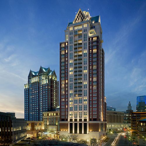 The image shows two tall, illuminated buildings in a city during dusk, with smaller buildings and light trails from cars in the foreground.