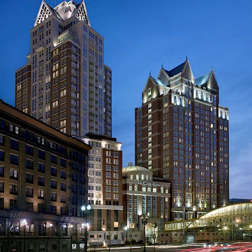 The image shows tall, illuminated buildings with gothic-style rooftops, captured against a twilight sky, alongside light streaks from moving vehicles.