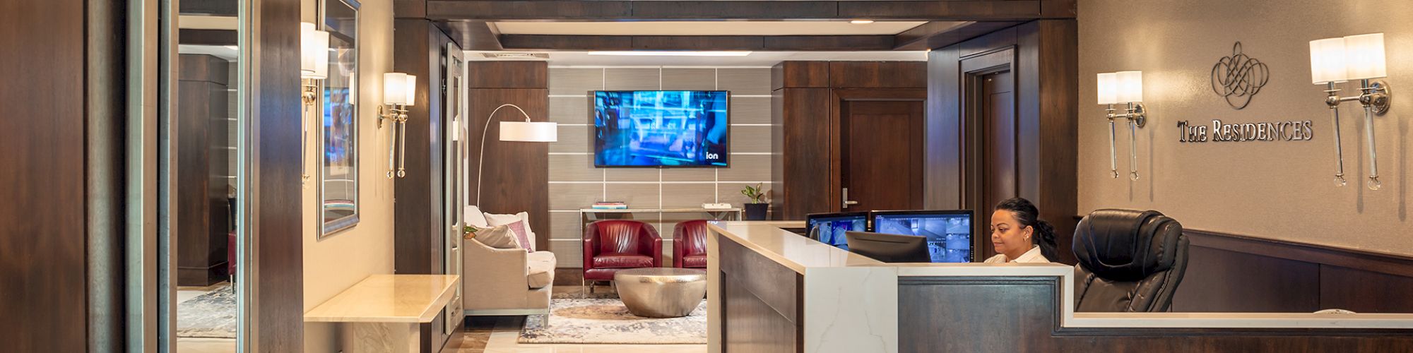 A modern reception area with a desk, a receptionist, wood paneling, wall-mounted lights, a TV, and a seating area in the background.