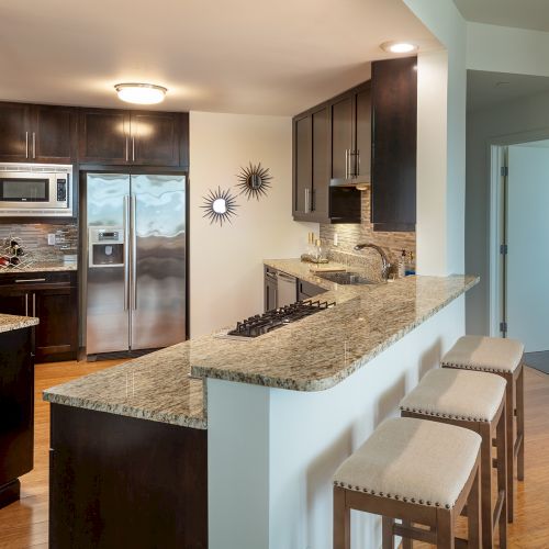 Modern kitchen with dark cabinets, stainless steel appliances, granite countertops, bar stools, and sunburst wall decor, adjoined by hallway.