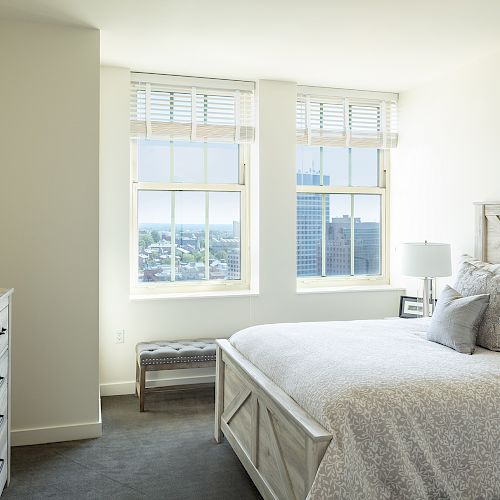 A bright bedroom with a large bed, two windows with city views, a dresser with decor, a bench below the windows, and minimalist decor on the walls.