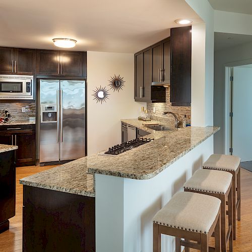 A modern kitchen with dark cabinets, stainless steel appliances, granite countertops, a bar area with stools, and decorative wall mirrors near an open doorway.