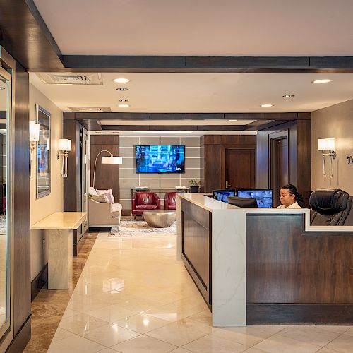 A modern reception area with a receptionist, desk, wall-mounted TV, and seating area. Warm lighting and wooden accents add to the ambiance.