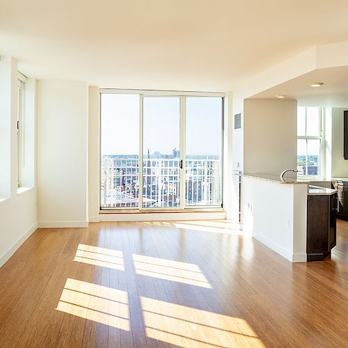 A bright, modern apartment with large windows, wooden flooring, a kitchen with dark cabinets and stainless steel appliances, and a small balcony.