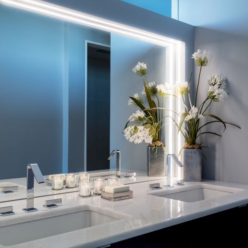 A modern bathroom with a large illuminated mirror, double sinks, a vase with white flowers, and several candles and toiletries on the counter.