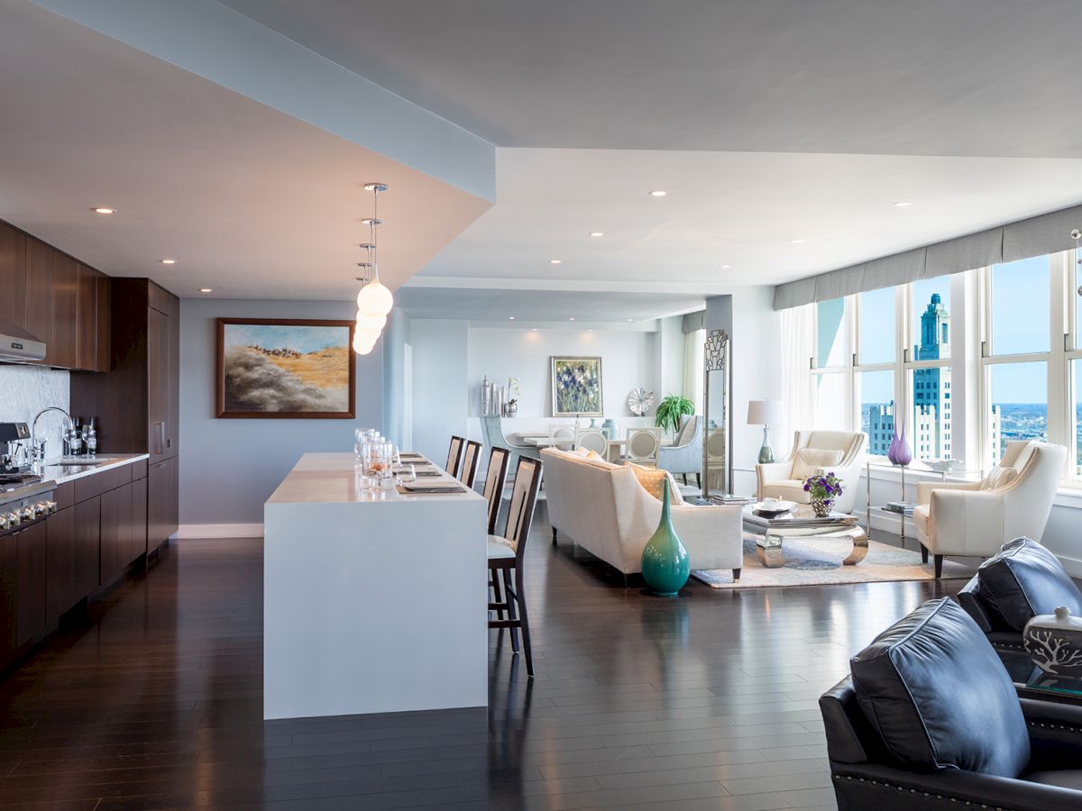 Modern open-plan kitchen and living area with sleek design, featuring a large island, dark wood flooring, contemporary furniture, and city views.